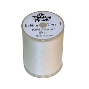 May include: A spool of white bobbin thread. The label reads "The Finishing Touch", "Bobbin Thread", "100% Polyester 60 wt." and "Made in Japan". The thread is tightly wound on a clear plastic spool.
