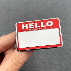 Peut inclure: Une épingle de badge rectangulaire avec un fond rouge et un bord argenté. L'épingle comporte le mot "HELLO" au-dessus du texte "MY NAME IS" et un espace blanc pour un nom.