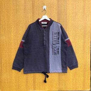 Puede incluir: Una sudadera azul oscuro y gris con ribetes burdeos. La sudadera tiene un cuello abotonado y un cordón en la parte inferior. El texto "NEXT TIME IS THE BEST WITH VIGOR" está impreso en el lado derecho de la sudadera.