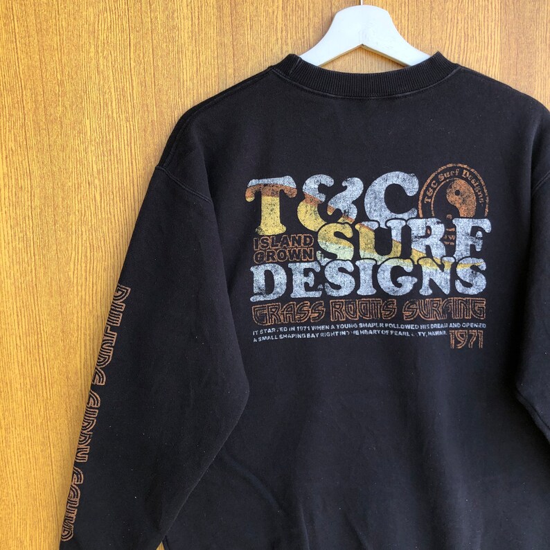 May include: Black sweatshirt with a graphic print on the back that says "T&C Surf Designs Grass Roots Surfing" and "Island Grown" in a vintage style font. The text includes the year "1971".