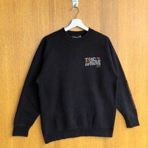 May include: Black long-sleeve sweatshirt with a brown and gold graphic that says "T&C Surf Designs 1971". The graphic is on the front of the sweatshirt. The sleeves have a brown and gold pattern.