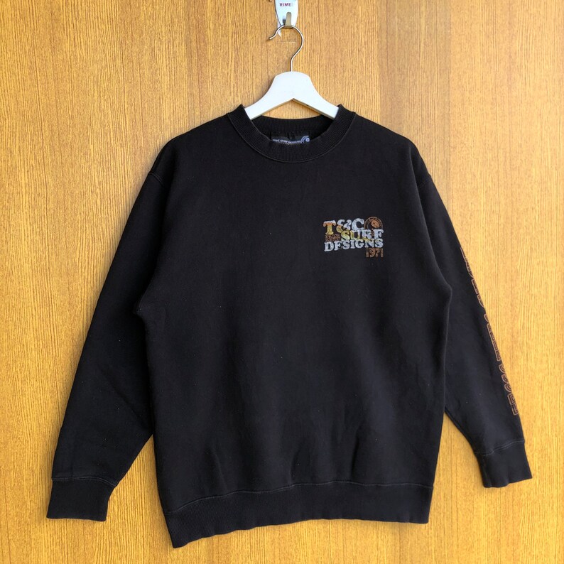 May include: Black long-sleeve sweatshirt with a brown and gold graphic that says "T&C Surf Designs 1971". The graphic is on the front of the sweatshirt. The sleeves have a brown and gold pattern.