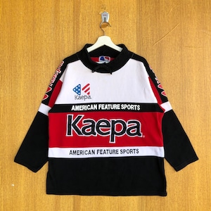 Op de afbeelding: Een zwart, rood en wit longsleeve sweatshirt met de tekst "American Feature Sports" en het logo "Kaepa" bedrukt op de voorkant. Het logo heeft een rood, wit en blauwe Amerikaanse vlag.