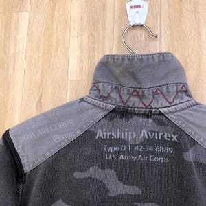 K&ouml;nnte beinhalten: Grau-schwarze Tarnjacke mit dem Text "Airship Avirex" und "U.S. Army Air Corps". Die Jacke hat einen grauen Kragen mit roten N&auml;hten und einen schwarzen Akzent auf der Schulter. Die Jacke h&auml;ngt an einem Metallb&uuml;gel.
