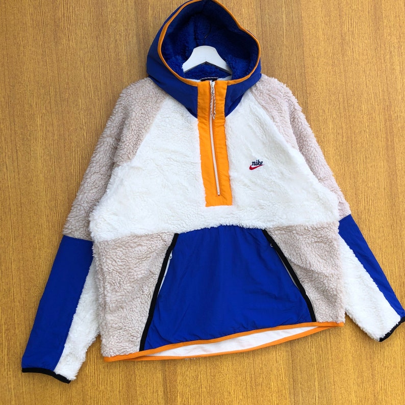 Puede incluir: Una sudadera con capucha de vell&oacute;n blanca, beige y azul con cierre de cremallera a la mitad y un logotipo de Nike en el pecho. La sudadera tiene una cremallera y ribetes de color naranja contrastantes.