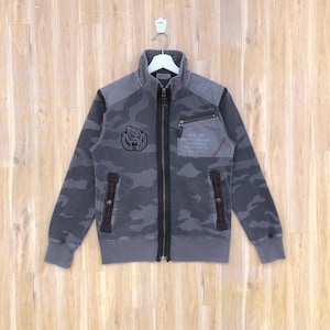 K&ouml;nnte beinhalten: Graue Camouflage-Rei&szlig;verschlussjacke mit hohem Kragen. Die Jacke hat eine kleine Brusttasche, einen Aufn&auml;her mit einem Emblem und rote Nahtdetails. Die Jacke h&auml;ngt an einem wei&szlig;en Kleiderb&uuml;gel vor einem Holzgrund.