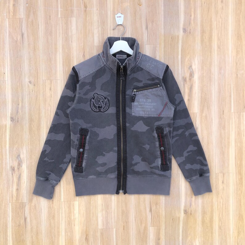 K&ouml;nnte beinhalten: Graue Camouflage-Rei&szlig;verschlussjacke mit hohem Kragen. Die Jacke hat eine kleine Brusttasche, einen Aufn&auml;her mit einem Emblem und rote Nahtdetails. Die Jacke h&auml;ngt an einem wei&szlig;en Kleiderb&uuml;gel vor einem Holzgrund.