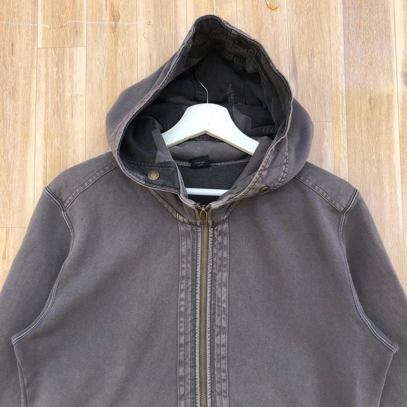 May include: A gray hooded zip-up sweatshirt with a drawstring hood and a full-length zipper. The sweatshirt has a worn, vintage look, with visible stitching details and a faded appearance. The hood is lined with a darker fabric.