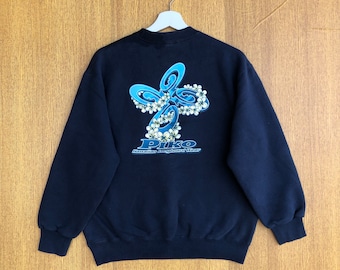 Può includere: Una felpa nera con una grafica blu e bianca di un disegno di fiori e il testo "PIKO Hawaiian Surfboard Wear" sul retro.