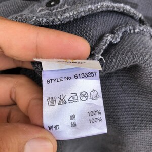 K&ouml;nnte beinhalten: Nahaufnahme eines grauen Stoffetiketts mit dem Text "STYLE No. 6133257". Das Etikett enth&auml;lt Waschsymbole und japanische Schriftzeichen. Der Stoff besteht aus 100 % Baumwolle.