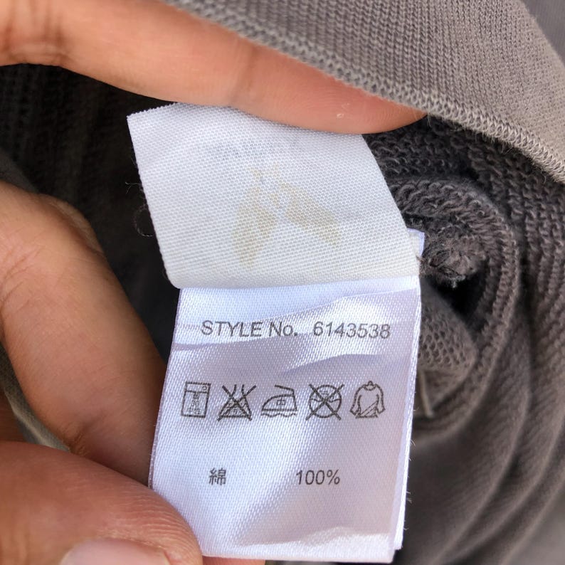 May include: A close-up of a clothing tag with the text "STYLE No. 6143538". The tag also includes care symbols and the text "綿 100%" in Japanese characters. The tag is attached to a gray garment.