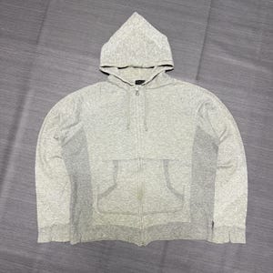 May include: A light gray heathered zip-up hoodie featuring a drawstring hood, long sleeves, and a split front kangaroo pocket. The garment is constructed with a central zipper closure and side panel detailing, showcasing a consistent textured knit fabric.