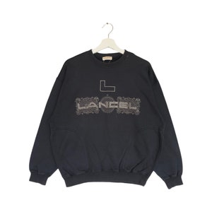 Könnte beinhalten: Ein schwarzes Sweatshirt mit dem Wort "LANCEL" in einem dekorativen Design auf der Brust. Das Sweatshirt hat einen Rundhalsausschnitt und lange Ärmel. Das Kleidungsstück hängt an einem weißen Kleiderbügel.