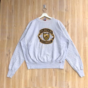 May include: Grey sweatshirt with a Purdue Boilermakers logo embroidered on the front. The logo is a shield with a gold outline and a black and white striped background. The text "Purdue Boilermakers" is written in black letters below the shield.
