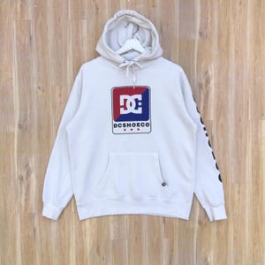 May include: White hoodie with a drawstring hood and a kangaroo pocket. The front features a logo with red, blue, and white colors, and the text "DCSHOECO". The sleeve has a black graphic. The hoodie is hanging on a wooden surface.