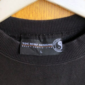 May include: Black T&C Surf Designs sweatshirt with a black tag that reads "T&C Surf Designs Enjoy Surfin' Life" with a white "L" size tag and a black and white yin yang symbol with the text "T&C Surf Hawaii" in orange.