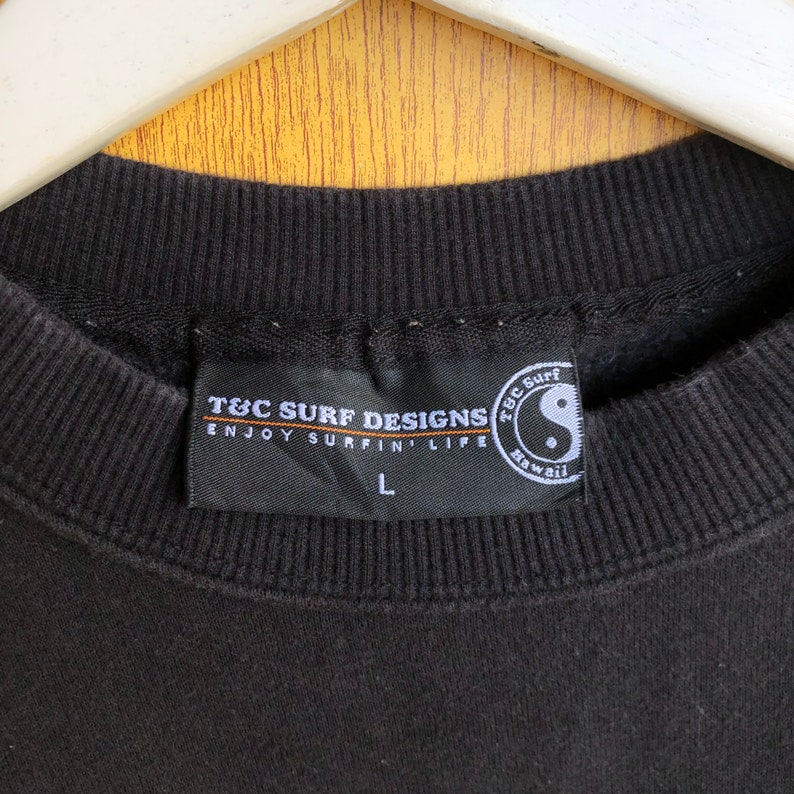 May include: Black T&C Surf Designs sweatshirt with a black tag that reads "T&C Surf Designs Enjoy Surfin' Life" with a white "L" size tag and a black and white yin yang symbol with the text "T&C Surf Hawaii" in orange.