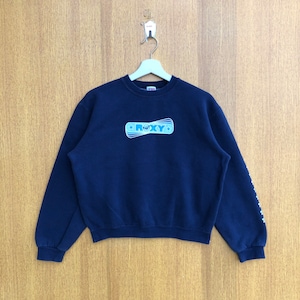May include: A navy blue sweatshirt with a white Roxy logo on the front. The logo is a stylised wave with the word Roxy written inside. The sweatshirt has a crew neck and long sleeves.