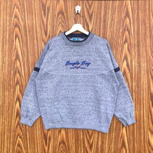May include: A gray sweatshirt with the text "Bugle Boy" embroidered in blue across the chest. The sweatshirt has a textured pattern, a navy blue collar, and navy blue stripes on the sleeves. The garment is hanging on a wooden background.