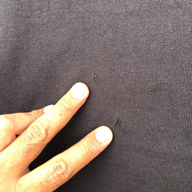 May include: Close-up of a dark gray fabric with a textured surface. The fabric has several small holes. A hand with visible skin tone and white fingernails is touching the fabric.