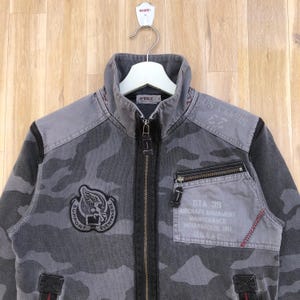 K&ouml;nnte beinhalten: Graue Tarnjacke mit durchgehendem Rei&szlig;verschluss. Die Jacke hat einen hohen Kragen, mehrere Taschen und Aufn&auml;her mit Text und Grafiken. Der Text enth&auml;lt "SUPPORT EVERYWHERE", "STA 39" und "USAAC".