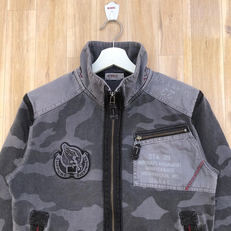 K&ouml;nnte beinhalten: Graue Tarnjacke mit durchgehendem Rei&szlig;verschluss. Die Jacke hat einen hohen Kragen, mehrere Taschen und Aufn&auml;her mit Text und Grafiken. Der Text enth&auml;lt "SUPPORT EVERYWHERE", "STA 39" und "USAAC".