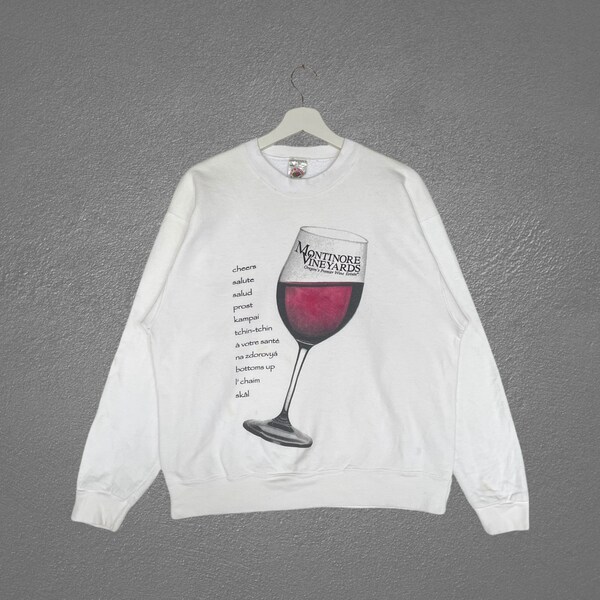 Könnte beinhalten: Weißer Langarm-Pullover mit einem Aufdruck eines Weinglases, gefüllt mit Rotwein. Auf dem Glas steht der Text "MONTINORE VINEYARDS". Der Text "cheere salute salud prost kampai tchin-tchin A votre santé na zdorovya bottons up Pchaim  العام" ist auf der linken Seite des Pullovers aufgedruckt.