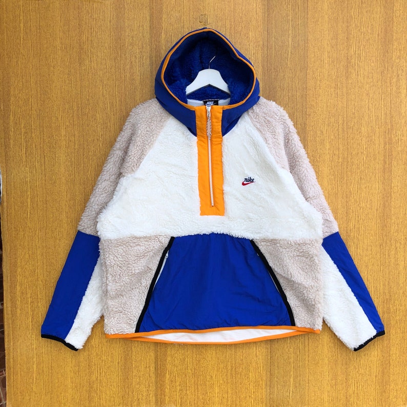 Puede incluir: Una chaqueta de forro polar blanca, beige y azul con cierre de cremallera a la mitad y un logotipo de Nike en el pecho. La chaqueta tiene capucha y un tirador de cremallera naranja contrastante.