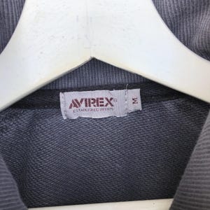 K&ouml;nnte beinhalten: Nahaufnahme eines grauen Sweatshirt-Etiketts mit dem Markennamen "AVIREX" in Rot und Wei&szlig; und dem Text "ESTABLISHED IN 1975". Das Etikett hat auch ein Gr&ouml;&szlig;enetikett "M".