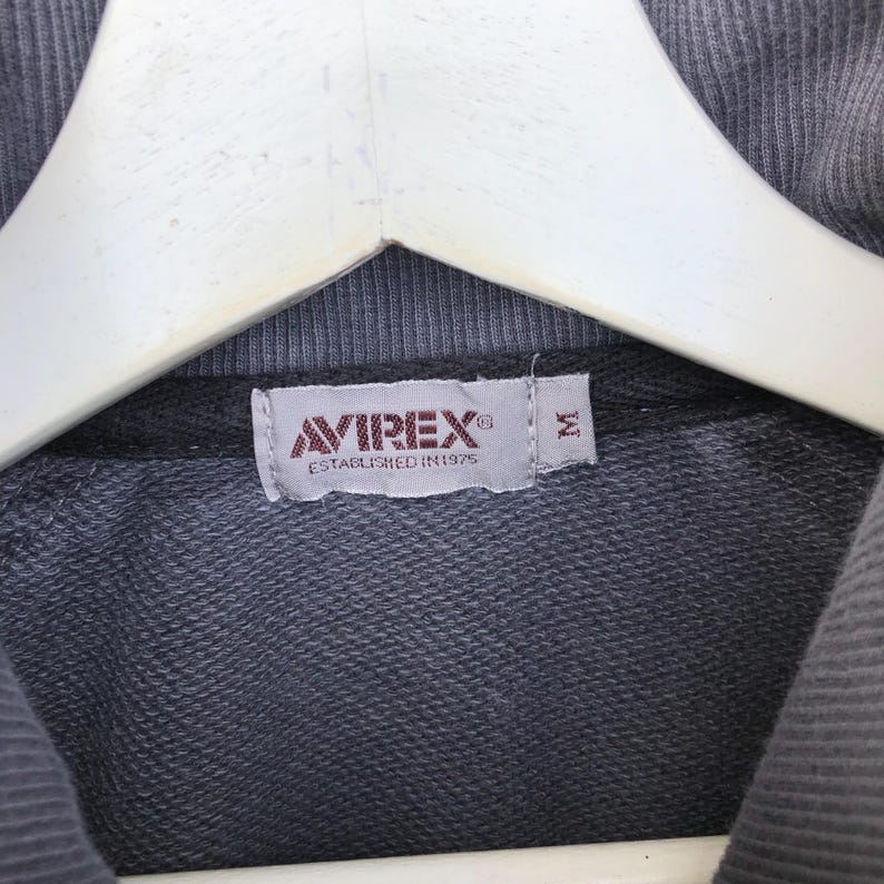K&ouml;nnte beinhalten: Nahaufnahme eines grauen Sweatshirt-Etiketts mit dem Markennamen "AVIREX" in Rot und Wei&szlig; und dem Text "ESTABLISHED IN 1975". Das Etikett hat auch ein Gr&ouml;&szlig;enetikett "M".