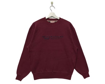 May include: A maroon-colored McGregor sweatshirt with long sleeves. The front features the brand name and logo in a dark blue embroidered design. The sweatshirt is hanging on a white hanger against a white background.