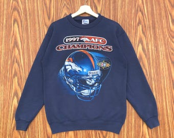 May include: Navy blue sweatshirt with the text "1997 AFC Champions" and a football helmet graphic. The design includes the Denver Broncos logo and a football. The sweatshirt is displayed against a wooden backdrop.