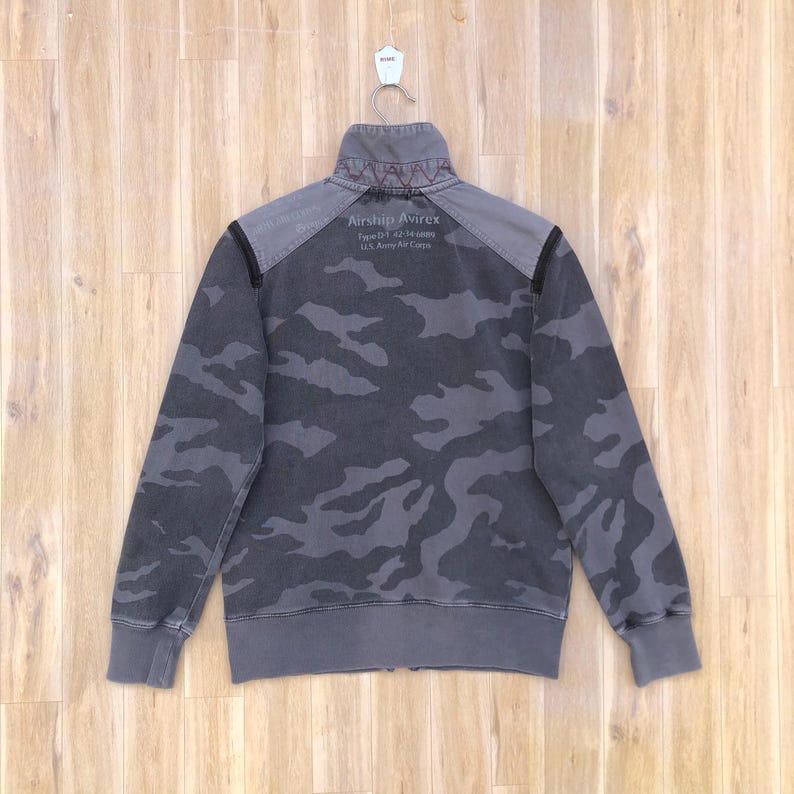K&ouml;nnte beinhalten: Graue Camouflage-Rei&szlig;verschlussjacke mit dem Text "Airship Avirex" auf dem oberen R&uuml;cken. Die Jacke hat einen hohen Kragen und gerippte B&uuml;ndchen und Saum. Die Jacke h&auml;ngt an einem wei&szlig;en Haken vor einem Holzgrund.