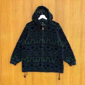 Può includere: Una giacca in pile con cappuccio blu scuro e verde con un motivo geometrico. La giacca ha una chiusura a zip e un cappuccio con coulisse.