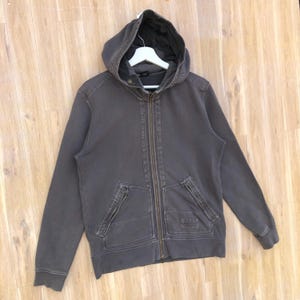 May include: A charcoal gray hooded sweatshirt with a full zipper closure. The sweatshirt has two front pockets with zippers and a small rectangular patch on the right pocket. The hood is lined with a darker gray fabric.