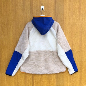 Puede incluir: Una chaqueta de forro polar blanca, beige y azul con capucha. La chaqueta tiene una cremallera y un ribete naranja contrastante alrededor de la capucha y los bolsillos.