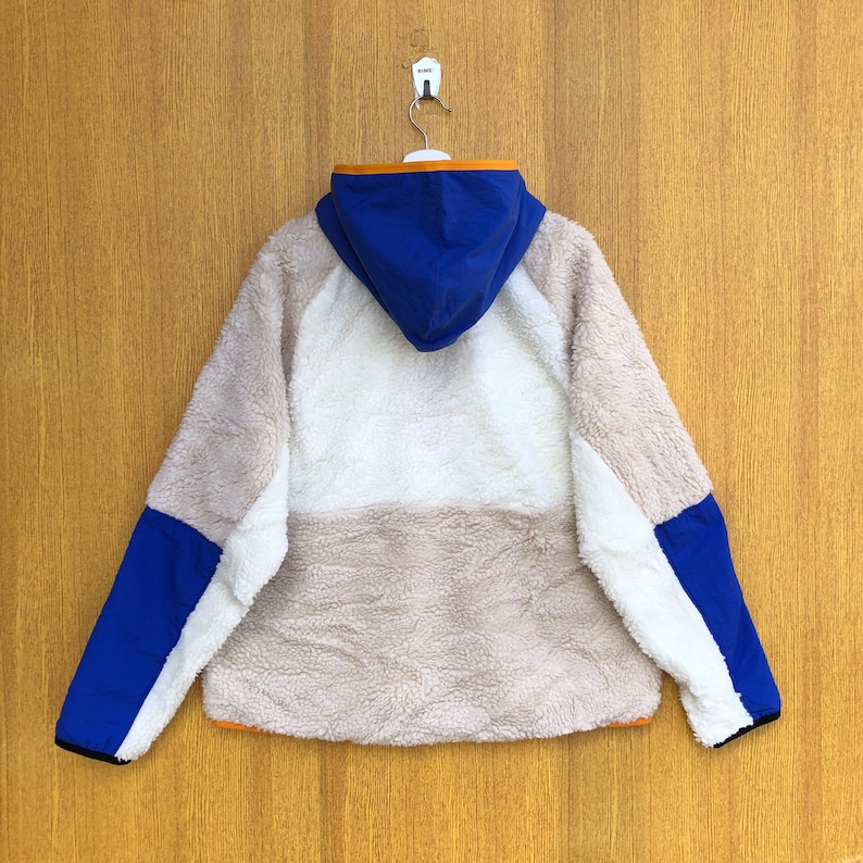 Puede incluir: Una chaqueta de forro polar blanca, beige y azul con capucha. La chaqueta tiene una cremallera y un ribete naranja contrastante alrededor de la capucha y los bolsillos.