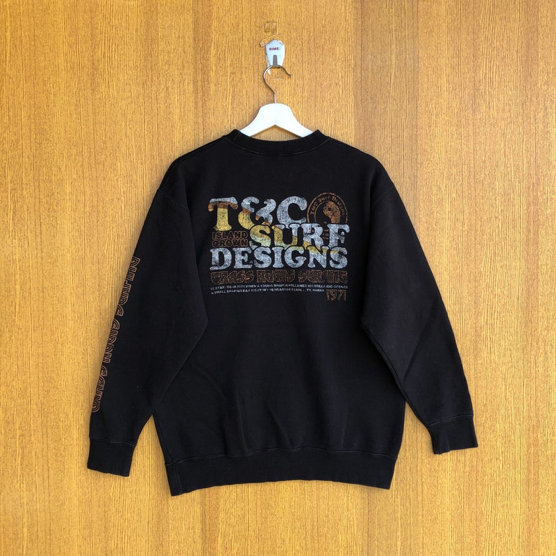 May include: Black sweatshirt with a brown and gold graphic print on the back that says "T&C Surf Designs Island Grown Grass Roots Surfing 1971". The graphic also includes a small circle with a bear inside. The sleeves have a brown and gold print that says "Island Grown" repeated.