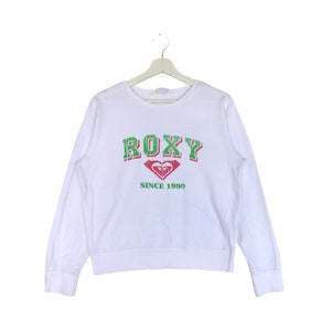 May include: White long-sleeved sweatshirt featuring the word "ROXY" in green and red block letters. Below the word "ROXY" is a heart-shaped logo and the text "SINCE 1990" in green.