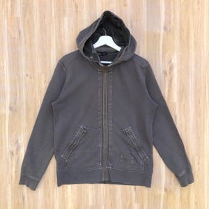May include: A gray zip-up hoodie with a hood, two front pockets with zippers, and a small logo on the lower right pocket. The hoodie is hanging on a white hanger against a light brown wooden background.