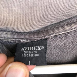 May include: Close-up of a gray garment tag with the brand name "AVIREX&reg;" in white letters. Below the brand name, the text "GUARANTEE GOOD FOR ONE" is printed. The tag also features the size "L".