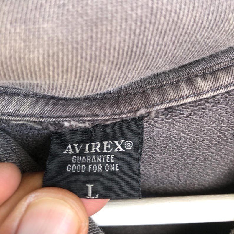 May include: Close-up of a gray garment tag with the brand name "AVIREX&reg;" in white letters. Below the brand name, the text "GUARANTEE GOOD FOR ONE" is printed. The tag also features the size "L".