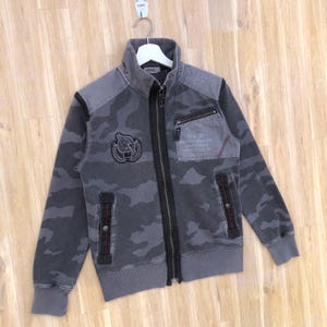 K&ouml;nnte beinhalten: Graue Camouflage-Rei&szlig;verschlussjacke mit hohem Kragen. Die Jacke hat einen Aufn&auml;her mit einem Adleremblem und Text, eine kleine Rei&szlig;verschlusstasche und rote Nahtdetails. Die Jacke h&auml;ngt an einem wei&szlig;en Kleiderb&uuml;gel.