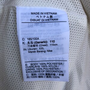 Puede incluir: Una etiqueta blanca con texto negro que detalla el tama&ntilde;o de la prenda, la composici&oacute;n del material y el origen. La etiqueta dice "MADE IN VIETNAM", "ベトナム製", "DIBUAT DI VIETNAM", "C: 185/100A", "K: 호칭 (Generic): 110", "가슴둘레 (Chest): 110cm", "IN: Ukuran: XXL", "Merek Barang: Nike", "E: BODY: 100% POLYESTER / OVERLAYS: 100% NYLON / LINING: 100% POLYESTER/ EXCLUSIVE OF DECORATION", "C: 面料:100% 聚酯纤维/外接 片:100%锦纶/里料:100%"