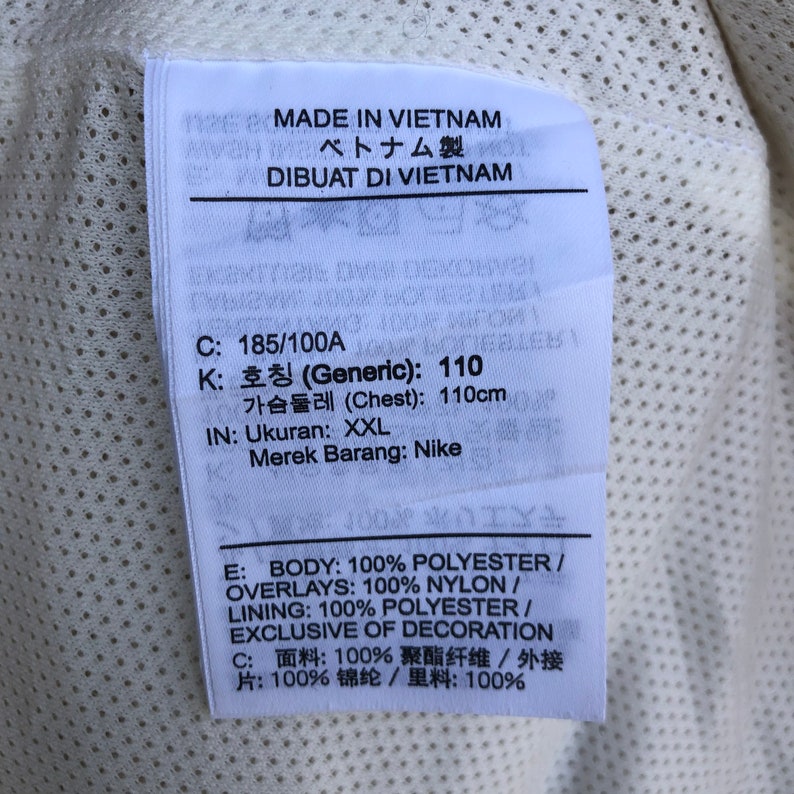 Puede incluir: Una etiqueta blanca con texto negro que detalla el tama&ntilde;o de la prenda, la composici&oacute;n del material y el origen. La etiqueta dice "MADE IN VIETNAM", "ベトナム製", "DIBUAT DI VIETNAM", "C: 185/100A", "K: 호칭 (Generic): 110", "가슴둘레 (Chest): 110cm", "IN: Ukuran: XXL", "Merek Barang: Nike", "E: BODY: 100% POLYESTER / OVERLAYS: 100% NYLON / LINING: 100% POLYESTER/ EXCLUSIVE OF DECORATION", "C: 面料:100% 聚酯纤维/外接 片:100%锦纶/里料:100%"