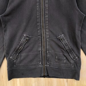 May include: Charcoal gray zip-up hoodie with two front pockets and a full-length zipper. The hoodie has a distressed, vintage look with visible stitching details and a small patch on the right pocket. The fabric appears to be a medium-weight cotton blend.
