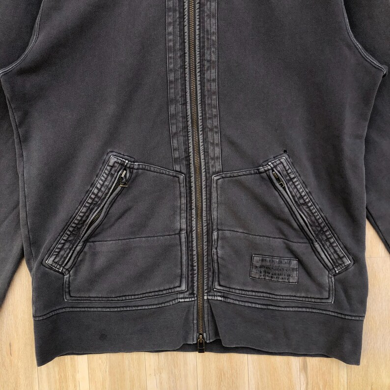May include: Charcoal gray zip-up hoodie with two front pockets and a full-length zipper. The hoodie has a distressed, vintage look with visible stitching details and a small patch on the right pocket. The fabric appears to be a medium-weight cotton blend.