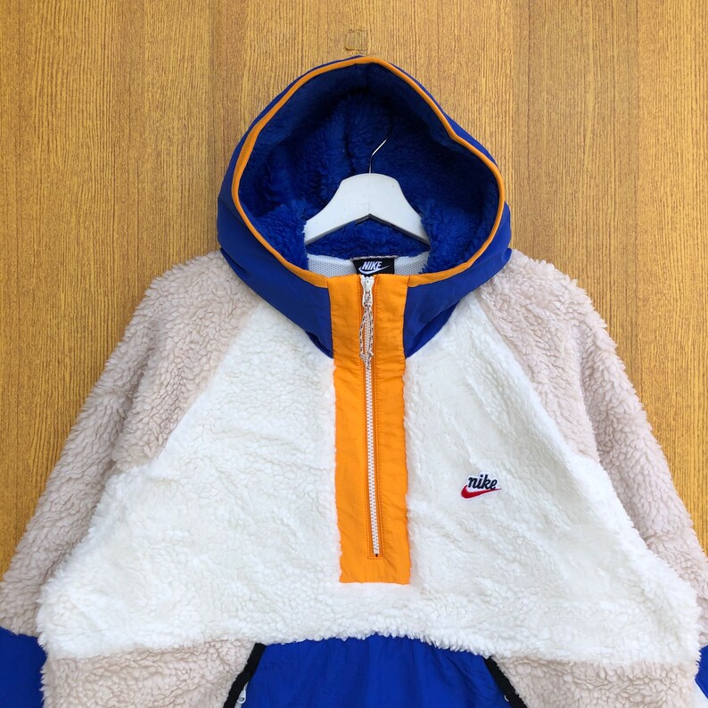 Puede incluir: Una chaqueta de forro polar con capucha azul y blanca con una cremallera naranja contrastante y un peque&ntilde;o logotipo "nike" rojo en el lado derecho del pecho.