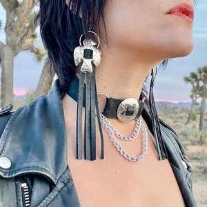 May include: A woman wearing a black leather jacket with a silver choker necklace featuring a large silver pendant and a chain. She is also wearing silver earrings with black tassels.