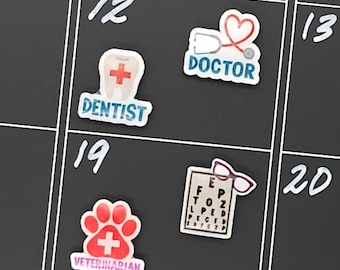 May include: A black calendar with white numbers and medical-themed magnets. Magnets include a tooth with a cross and the word "DENTIST", a stethoscope with a heart and the word "DOCTOR", a paw print with a cross and the word "VETERINARIAN", and an eye chart with glasses.